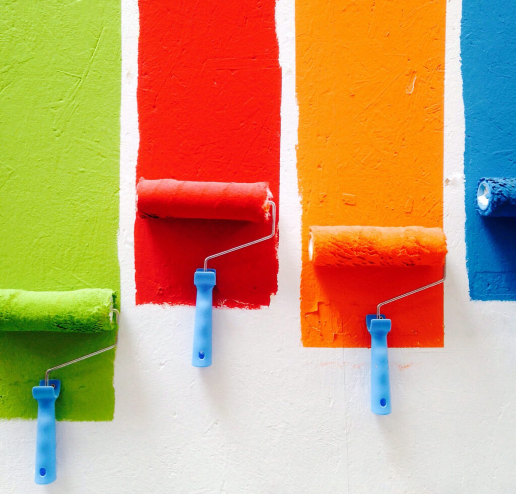 Bilde viser fire maleruller som maler en hvit vegg, Malerkostene maler en stripe av hver av fargene grønn, rød, oransje og blå . 