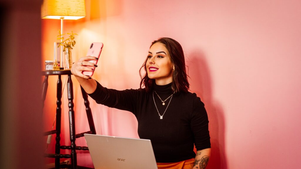 Bildet viser ei kvinne som sitter foran en laptop. Hun tar en selfie med en rosa mobiltelefon.